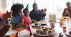 一个戴着派对帽的家庭围坐在桌子旁，为一个笑着的生日男孩鼓掌。