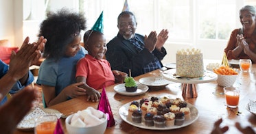 A family wearing party hats sits around a table, clapping for a laughing birthday boy who is about t...