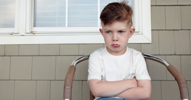 A boy crosses his arm and pouts sitting in a chair outside his house.