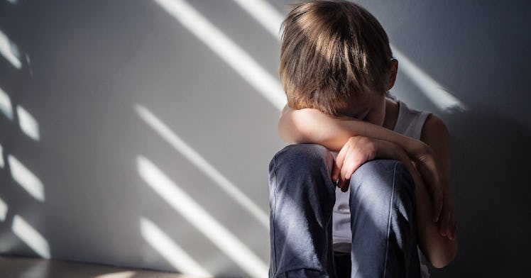 A child sits against a wall in a dark room with his head buried in his arm.