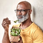A man sits against a wall, eating a salad.
