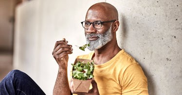 A man sits against a wall, eating a salad.