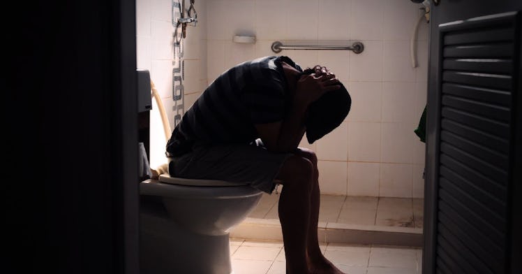 A man in a dark bathroom sits hunched over on the toilet.