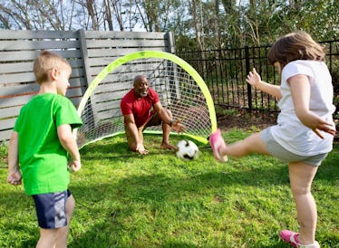 A dad and two kids playing soccer outdoors with a tiny soccer net behind them