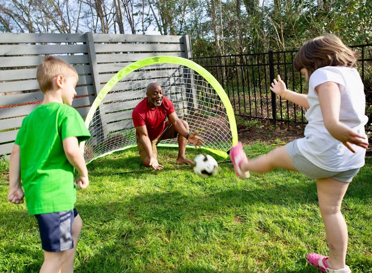 A dad and two kids playing soccer outdoors with a tiny soccer net behind them