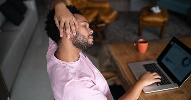 A man sits in a chair with perfect posture, stretching his neck while working on his laptop.