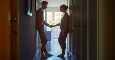 a husband and wife hold hands loosely, contemplating divorce, in a hallway backlit by a sunny window