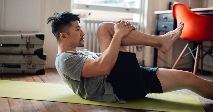 A man stretches on his back on a yoga mat at home, pulling his knee to his chest.