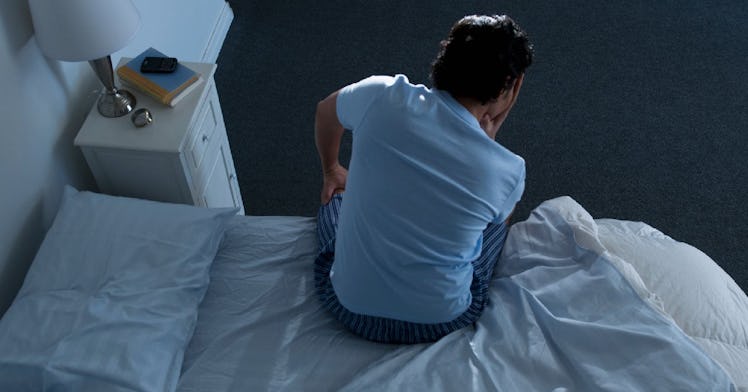 A man in pajamas sits on the edge of his bed sadly, facing away.