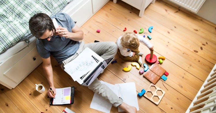 a dad sits on a bedroom floor next to his baby, working