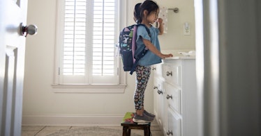 a little girl with a backpack on brushes her teeth in front of a bathroom mirror