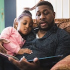 A dad reads a book to his daughter while they sit on a chair.