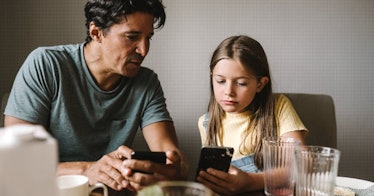 A dad and daughter sit at the dinner table, discussing something they see on their smart phones.