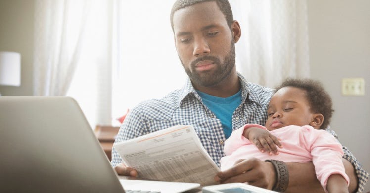 a dad looks at a bill with his infant sleeping in his arms