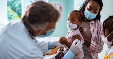 A toddler receives the COVID vaccine in a doctor's office, surrounded by her mother and sister.