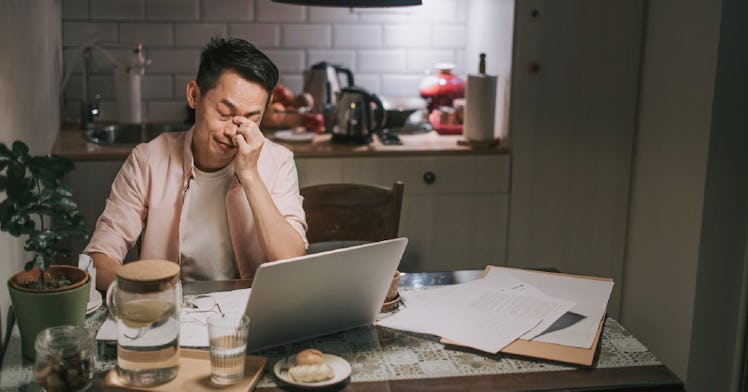 A man with brain fog does work at a laptop in his kitchen, looking frustrated.