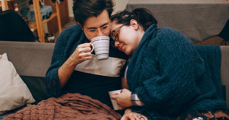 A couple with the flu is holed up together at home, wrapped in blankets and drinking tea.