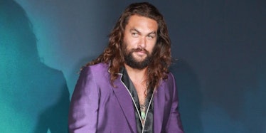 Jason Momoa in an unbuttoned black shirt and a purple blazer in front of a blue background