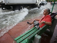 An older man sitting on a green bench next to sea while a wave is almost reaching him 