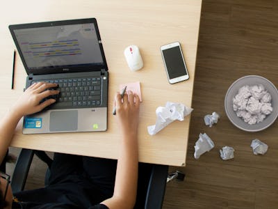 A woman typing on her laptop with a bin full of paper next to her