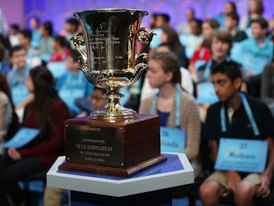 A close-up of the Scripps National Spelling Bee trophy with an audience blurred in the background