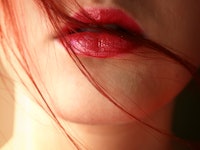 A close-up of the mouth of a red-haired woman with red lip gloss and strands of hair over her mouth