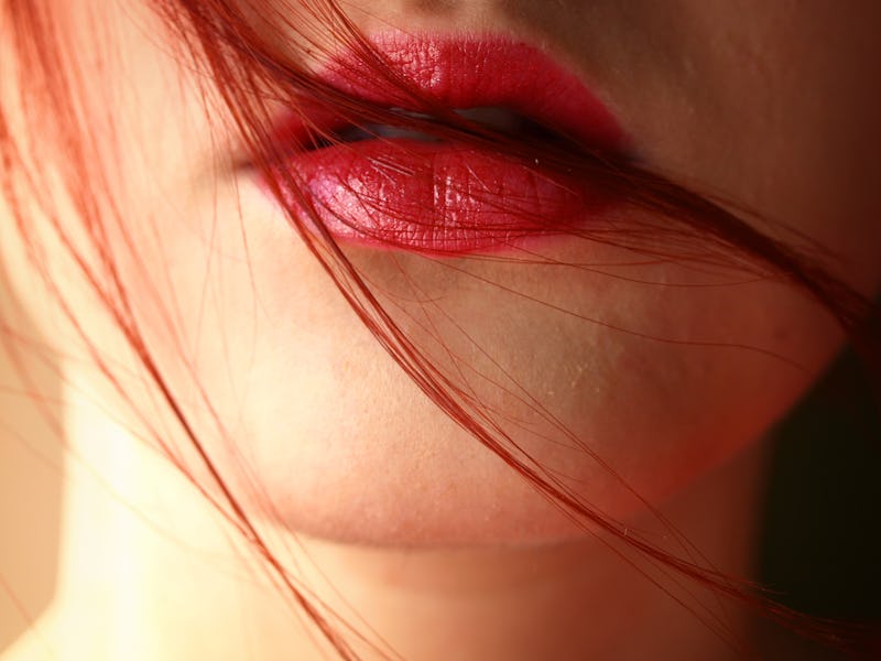 A close-up of the mouth of a red-haired woman with red lip gloss and strands of hair over her mouth