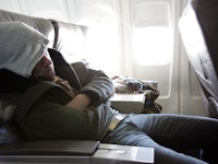 A man in a grey hoodie sleeping in an airplane seat and experiencing jet lag