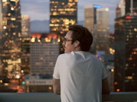 A man leaning onto a terrace fence with blurred lights of a city in the background