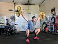 A man in a grey shirt, black short, and black cap squatting and holding a heavy weight over his body