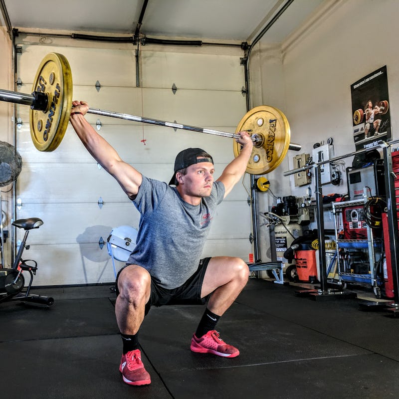 A man in a grey shirt, black short, and black cap squatting and holding a heavy weight over his body