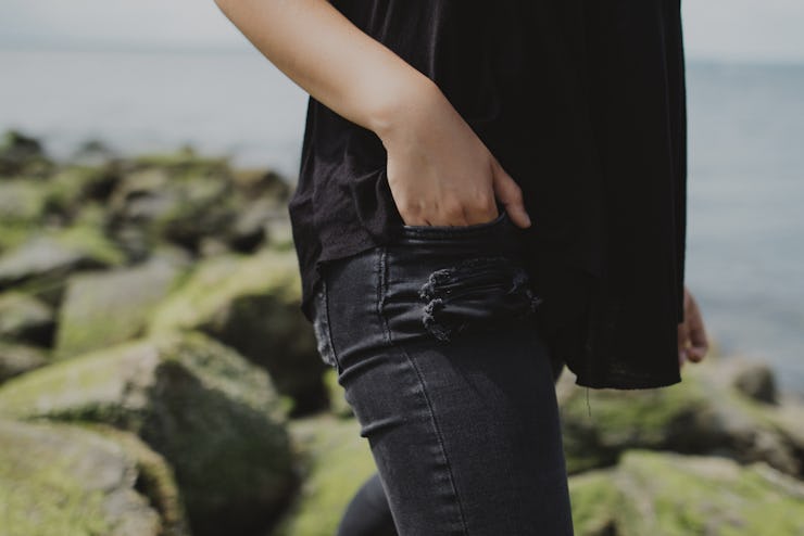 A woman putting her hand in her small pocket while standing on rocks next to a body of water