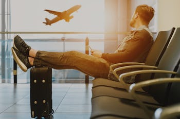 Younger male sitting on an airport chair with legs leaned on his suitcase