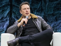 Elon musk in a brown leather jacket, black shirt, and black pants holding a microphone during an int...