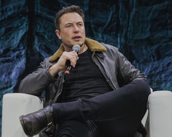 Elon musk in a brown leather jacket, black shirt, and black pants holding a microphone during an int...