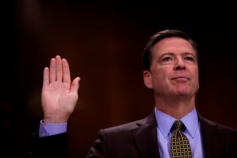 James Comey in a black suit, and blue shirt holding a hand up with an open palm