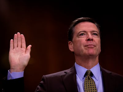 James Comey in a black suit, and blue shirt holding a hand up with an open palm