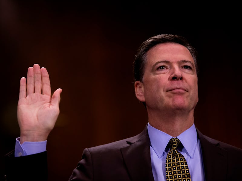 James Comey in a black suit, and blue shirt holding a hand up with an open palm