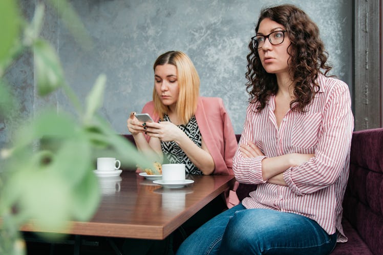 Two girls sitting at a caffe bar, where one is angry while the other is using her phone