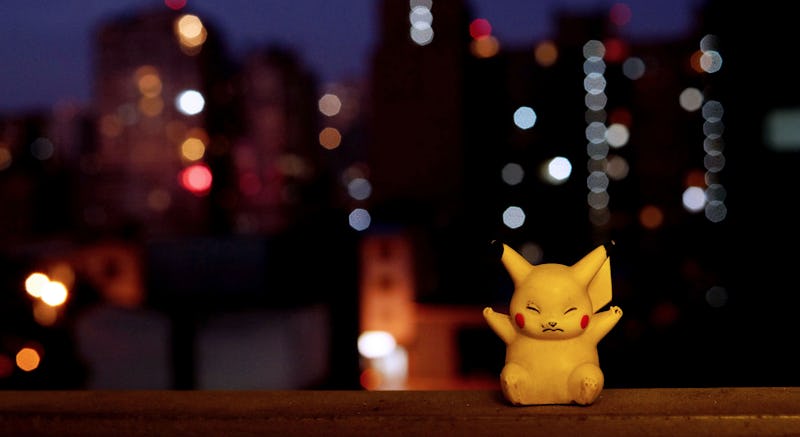 A tiny Pikachu from Pokemon Go toy next to a window with blurry city lights in the background.