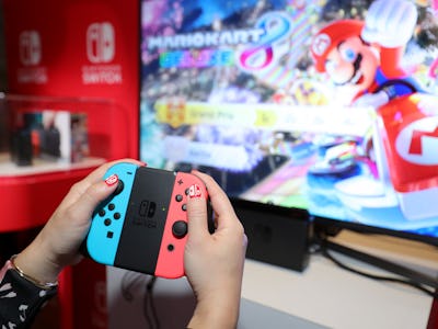 A person using a red and blue controller for Nintedno Switch with a large PC monitor in the backgrou...