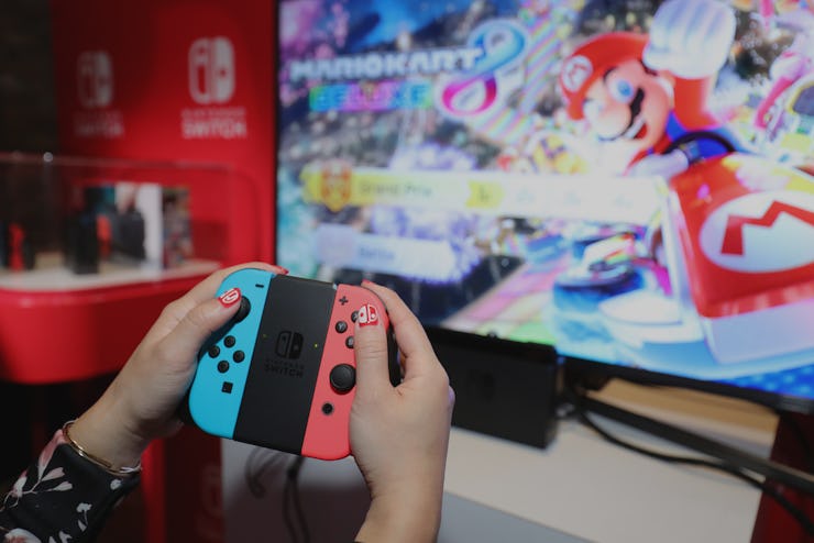 A person using a red and blue controller for Nintedno Switch with a large PC monitor in the backgrou...