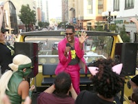 Orlando Jones in a pink suit holding a microphone while performing on the rooftop of an open bus