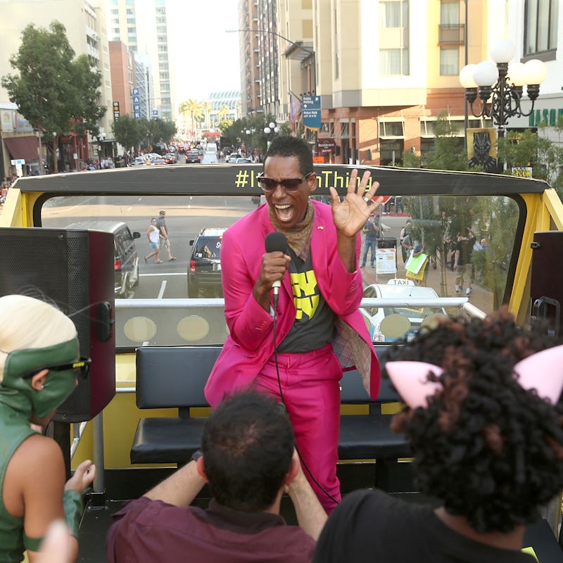 Orlando Jones in a pink suit holding a microphone while performing on the rooftop of an open bus