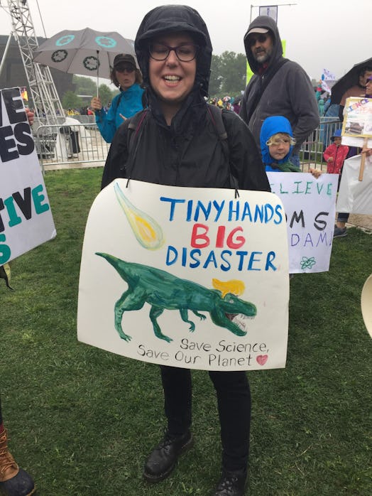 A woman holding a poster with "tiny hands big disaster" text and a dinosaur drawing