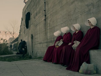 Handmaids from the Handmaids tale sit next to a large wall, dressed in their traditional uniforms.