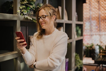 A woman leaning against a book shelf and scrolling through an online dating app on her phone
