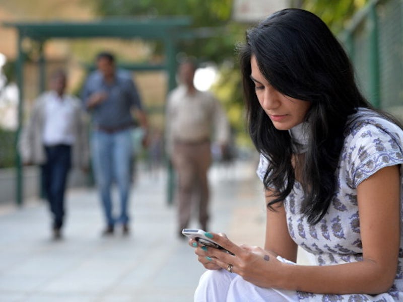A brunette woman sitting on a bench in a park and using a cellphone that is linked to cancer accordi...