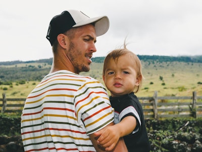 A dad in a striped shirt and a cap holding his toddler in his arms