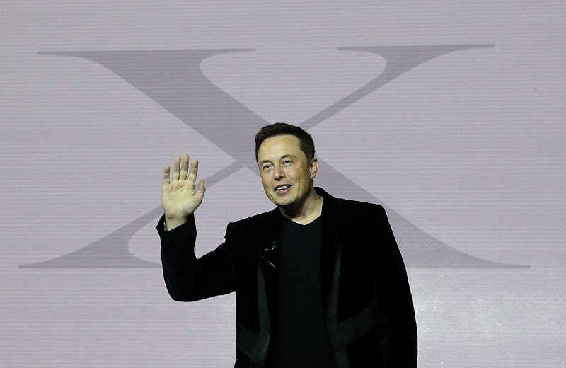 Elon Musk in a black suit waving with his right hand and a large 'X' on a white wall behind him
